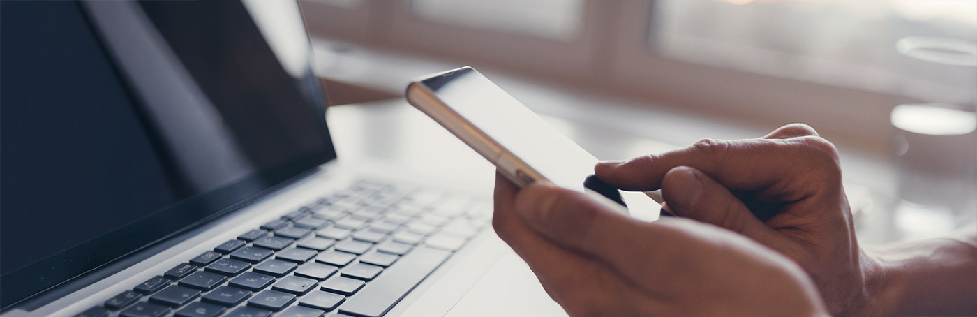 A close-up of a person's hands as the search on their phone, with a laptop on the desk in front of them