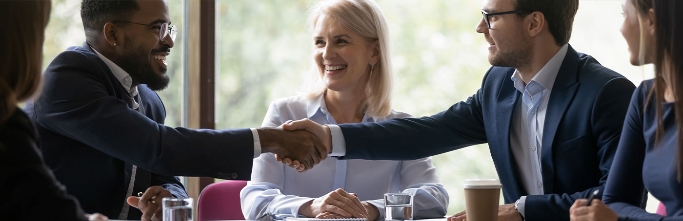 Two men in business suits shake hands in front of a woman in a meeting