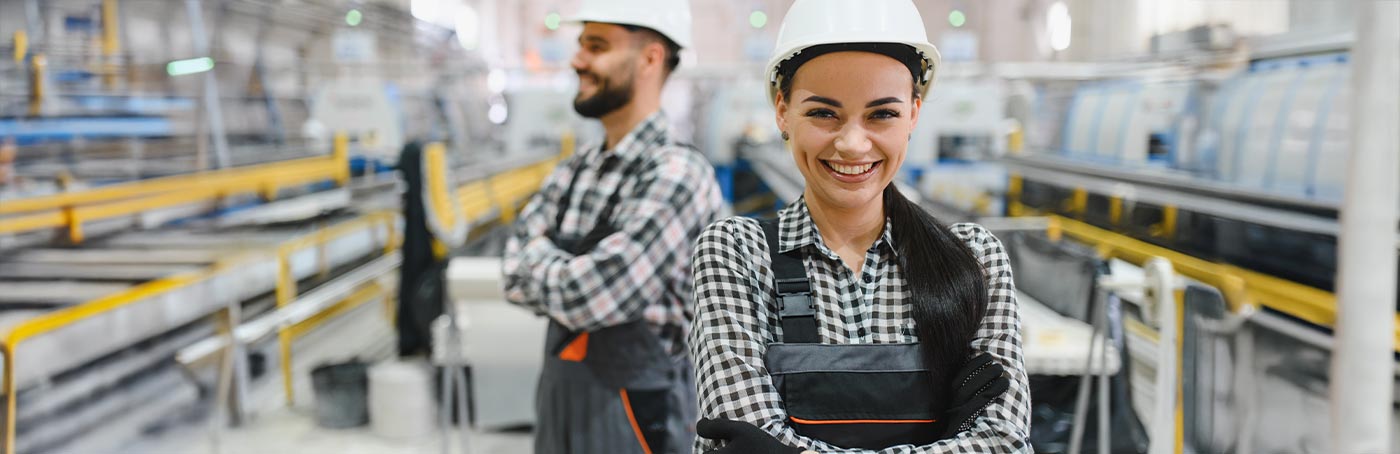 A woman employee in a hard hat smiling on the floor of a warehouse, with a smiling male co-worker behind her